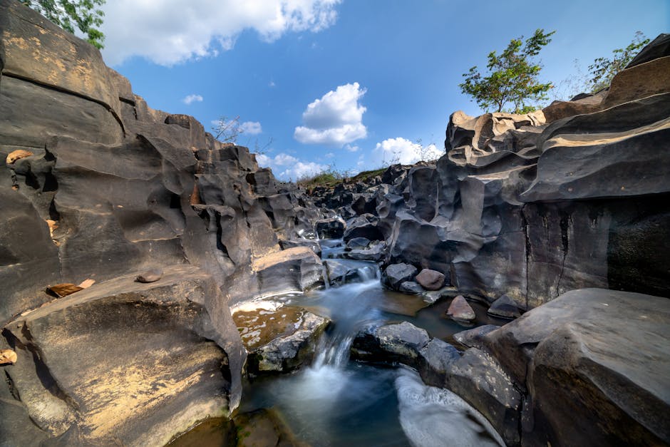 A natural rocky landscape featuring a narrow stream of water flowing between large, dark volcanic rocks with rugged textures and smooth, weathered surfaces. The rocks, varying in shades of black, grey, and brown, form steep, irregular walls on either side of the watercourse, with some boulders partially submerged in the shallow, clear stream. In the background, a small tree with green foliage extends branches over the rocks, contrasted against a bright blue sky dotted with fluffy white clouds, suggesting a sunny day. The environment appears undisturbed and natural, with no visible human intervention or equipment, embodying an untouched outdoor scene that could be part of a remote natural reserve or rural landscape. The scene's lighting highlights the textures and contours of the rocks, creating a visually appealing and tranquil setting suitable for depicting natural formations in a waste management or environmental context.