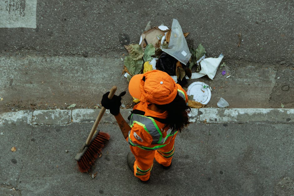 An individual wearing an orange high-visibility jacket and matching orange hat is seen from above, standing on a paved sidewalk next to a concrete curb. The person is holding a long-handled broom with a wooden handle and red bristles, and is in the process of sweeping scattered rubbish along the edge of the pavement. The rubbish includes crumpled paper, plastic bags, packaging, and food containers, some of which are partially crushed or torn. The rubbish pile is situated against a weathered, grey concrete or brick wall with visible texture and minor discoloration. The environment appears to be an outdoor urban setting, possibly a street or alleyway, with natural light illuminating the scene evenly. The presence of personal safety gear and the act of clearing debris suggests a private waste clearance or rubbish removal service aimed at maintaining cleanliness and proper waste disposal in the area, aligning with the service offerings of Waste Clearance Maida Vale.