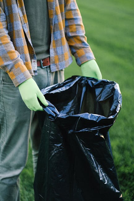 A person standing outdoors on a grassy area, wearing a plaid shirt over a green t-shirt, light brown trousers, and bright green gloves, holding open a large black rubbish bag made of glossy plastic. The individual appears to be preparing for waste collection or clearing, with the bag partially filled and positioned in front of them. The background shows a lush, green outdoor environment, suggesting a garden or park setting. This scene exemplifies private waste management practices, such as independent rubbish removal or garden waste disposal, often handled by services like Waste Clearance Maida Vale to aid in efficient rubbish collection and on-site clearance tasks. The image emphasizes the preparation phase of rubbish removal, highlighting the use of professional-grade, durable black bin bags suitable for collecting garden or general waste during an alternative waste handling process.
