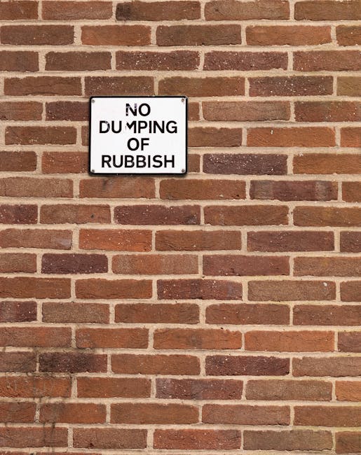 A rectangular white metal sign with black text reading 'NO DUMPING OF RUBBISH' is mounted on a brick wall. The wall consists of uniformly-sized reddish-brown bricks with a slightly rough texture and visible mortar joints, arranged in a traditional horizontal pattern. The sign is positioned towards the upper left portion of the image, with the brick wall extending both above and below it. The lighting is even, illuminating the brick surface clearly. This scene may relate indirectly to waste management regulations or private rubbish collection services, emphasizing controlled disposal through visible signage. The overall environment suggests an external exterior wall, part of a building’s façade or boundary wall, in an urban setting where managing rubbish is essential for cleanliness and compliance with local waste policies.