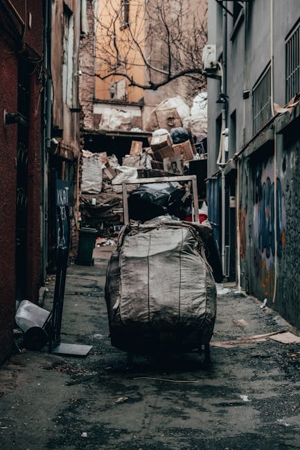 A narrow urban alleyway filled with a large pile of mixed waste and discarded packaging, with several cardboard boxes and black and white plastic bags stacked haphazardly at the end of the alley. In the foreground, a grey, weathered trolley bag with handles and visible stitching is positioned centrally on the asphalt surface, which is stained and dusty. The alley is flanked by dark, graffitied walls on either side, with some bare tree branches extending overhead from the background, where older brick buildings are visible above the clutter. The scene appears to be an example of private waste collection or rubbish removal in an urban setting, with Waste Clearance Maida Vale possibly providing a service for clearing such accumulated waste from similar alleyways. The lighting is diffuse, suggesting overcast conditions, and the overall atmosphere indicates a temporary or ongoing rubbish clearance process typical of independent waste disposal activities in busy city environments.