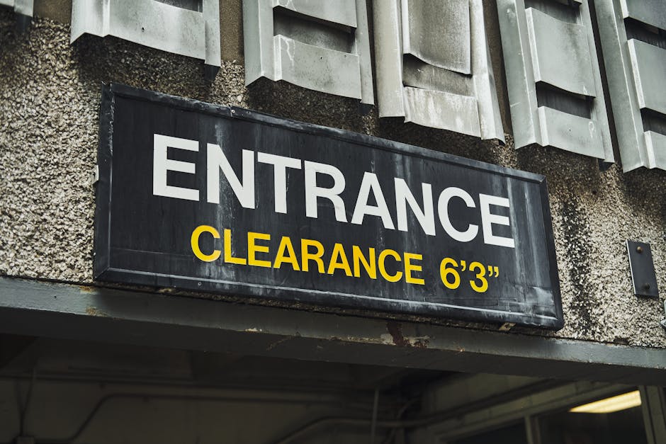 A black rectangular sign with white and yellow lettering reading 'ENTRANCE' and 'CLEARANCE 6’3”', mounted on a textured, weathered concrete building exterior. Above the sign, there are multiple grey, rectangular, vent-like fixtures with a ribbed surface, arranged in a horizontal row along the wall. The wall surface shows signs of age and wear, with visible patches of discoloration and rough texture. The lighting appears to be natural, highlighting the textures and contrasting colors of the sign and building materials. This setting suggests an entrance to a parking garage or service area with specific height restrictions, relevant in the context of private waste and rubbish removal access for waste clearance services like Waste Clearance Maida Vale.