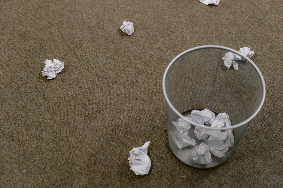 A clear, cylindrical waste paper bin made of semi-transparent plastic with a smooth, shiny surface is positioned on a brown textured carpeted floor. The bin contains several crumpled white sheets of paper, some of which are visibly spilling over the rim. Additional crumpled paper pieces are scattered on the surrounding carpet, with a few lying on the floor near the bin, indicating a recent or ongoing process of paper waste disposal. The setting appears to be an indoor environment, and the scene reflects a typical office or domestic space where informal rubbish collection or on-site waste disposal might occur, as managed by a service specializing in rubbish removal. The lighting is even and neutral, highlighting the texture of the carpet and the crumpled paper's irregular surface, contributing to a straightforward, factual presentation aligned with professional waste clearance activities.