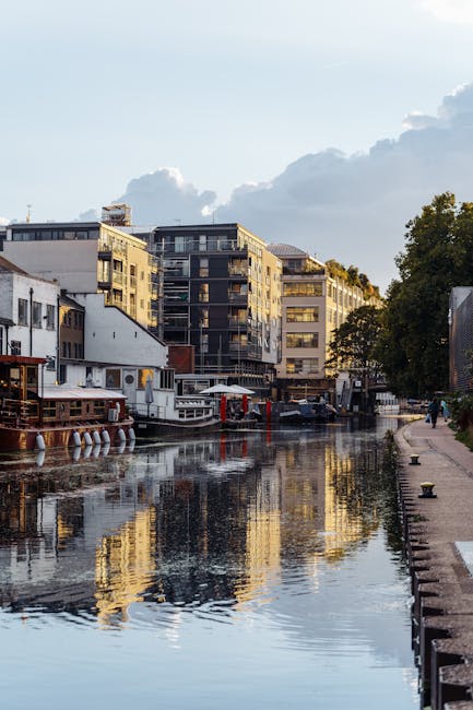 A daytime view of a narrow canal in Little Venice, Maida Vale, showing several modern multi-storey residential buildings with glass balconies and light-colored facades lining the water's edge. The canal's calm surface reflects the buildings and the partly cloudy sky overhead. On the right side, a pedestrian walkway runs parallel to the canal, bordered by a low concrete edge and some trees providing greenery. To the left, moored boats and houseboats with visible details such as wooden exteriors, white and red painted sections, and nautical equipment are docked along the waterway. The scene captures a quiet, urban waterside environment, typical of independent or private waste disposal and rubbish removal activities occurring in this area, with no visible waste or debris present in the scene itself. The natural lighting suggests late afternoon or early evening, highlighting the textures of the buildings, boats, and water reflections, creating a peaceful atmosphere for residents and visitors alike.