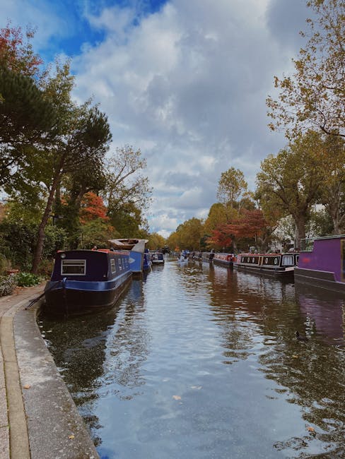 A daytime view of a narrow canal in Little Venice, Maida Vale, showing several modern multi-storey residential buildings with glass balconies and light-colored facades lining the water's edge. The canal's calm surface reflects the buildings and the partly cloudy sky overhead. On the right side, a pedestrian walkway runs parallel to the canal, bordered by a low concrete edge and some trees providing greenery. To the left, moored boats and houseboats with visible details such as wooden exteriors, white and red painted sections, and nautical equipment are docked along the waterway. The scene captures a quiet, urban waterside environment, typical of independent or private waste disposal and rubbish removal activities occurring in this area, with no visible waste or debris present in the scene itself. The natural lighting suggests late afternoon or early evening, highlighting the textures of the buildings, boats, and water reflections, creating a peaceful atmosphere for residents and visitors alike.