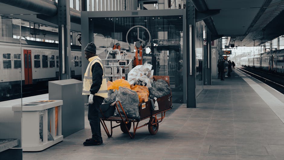 A worker dressed in dark clothing, including a beanie and gloves, stands beside a red hand-pulled trolley filled with large black, orange, and white plastic rubbish bags, as well as loose discarded materials, all positioned on a modern train station platform. The trolley is loaded with waste materials and is situated near a white recycling bin on the left. Behind the worker, there are metal and glass structures, industrial machinery, and a glass-enclosed lift, with train tracks visible to the right side of the platform. The scene is illuminated by natural daylight, highlighting the clean, tiled surface of the platform and the surrounding station environment. This setting reflects a disposal or rubbish collection process, consistent with private waste handling services such as those offered by Waste Clearance Maida Vale, within a contemporary urban transportation hub.