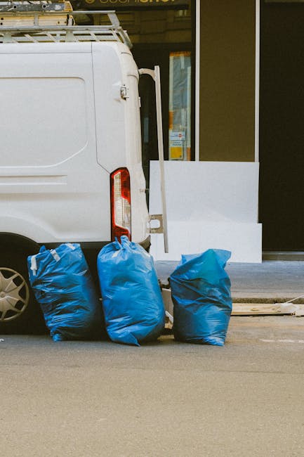 A worker dressed in dark clothing, including a beanie and gloves, stands beside a red hand-pulled trolley filled with large black, orange, and white plastic rubbish bags, as well as loose discarded materials, all positioned on a modern train station platform. The trolley is loaded with waste materials and is situated near a white recycling bin on the left. Behind the worker, there are metal and glass structures, industrial machinery, and a glass-enclosed lift, with train tracks visible to the right side of the platform. The scene is illuminated by natural daylight, highlighting the clean, tiled surface of the platform and the surrounding station environment. This setting reflects a disposal or rubbish collection process, consistent with private waste handling services such as those offered by Waste Clearance Maida Vale, within a contemporary urban transportation hub.