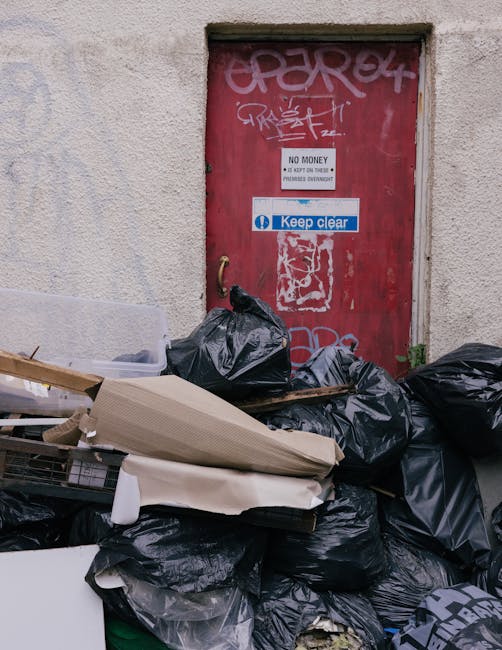 A pile of black rubbish bags filled with discarded waste stacked in front of a red metal door on a textured, off-white building wall. The bags appear full and are arranged haphazardly, with some leaning against the door and others on the ground. The door features multiple graffiti tags in white and pink spray paint, along with two rectangular signs: one indicating 'NO MONEY' and another blue and white sign reading 'Keep clear'. Surrounding the rubbish is a small section of pavement or driveway with minimal environmental features visible, suggesting an urban or residential alleyway setting. The scene depicts an instance of private waste disposal, typical of independent rubbish collection services, such as those offered by Waste Clearance Maida Vale, highlighting the need for proper rubbish removal in the area.