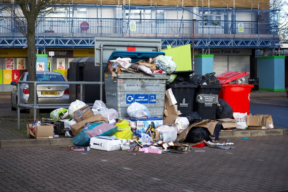 A large outdoor waste disposal area shows an overflowing collection of various rubbish items, including black bin bags, flattened cardboard boxes, and miscellaneous packaging materials, some spilling onto the paved ground. At the centre, a grey mixed paper and cardboard recycling bin is open, with its lid tilted back, revealing piled paper waste, such as newspapers, cardboard sheets, and paper bags. To the right, black waste bins are visible, one of which is filled with black plastic trash bags, while another red bin adjacent to them appears to be for general waste. A smaller white cardboard box and additional loose packaging are scattered at the front, with some items overlapping on the paved surface. Behind this scene, a blue and yellow commercial building with storefront signage and large windows is partially obscured by two parked cars—one grey on the left side of the image and another dark-colored. The background features a metal fence and an upper-floor balcony or scaffolding structure, suggesting ongoing building work or maintenance. The overall scene depicts a high volume of discarded waste awaiting collection, typical of private rubbish removal or alternative waste handling arrangements outside standard council services, with mild natural lighting indicating an overcast day.