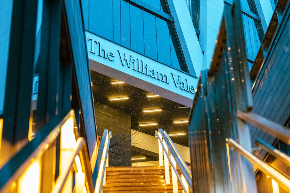 A modern multi-storey building with a blue exterior and illuminated signage reading 'The William Vale' is seen from below, with metal railings and a staircase leading up to the entrance. The scene is captured during the evening, with warm yellow lighting emanating from the building's interior and reflecting on the dark, rain-dotted glass surfaces. The surrounding environment includes parts of adjacent dark blue panels and a glimpse of the stairway's wooden steps. The overall atmosphere conveys a contemporary architectural style, emphasizing clean lines and sleek finishes, consistent with an urban hotel or venue. Waste Clearance Maida Vale’s rubbish removal services could be pertinent for clearing debris or renovation waste related to such modern commercial properties, especially during refurbishment or post-construction cleanup. This scene visually supports the importance of professional waste management for maintaining the cleanliness and functionality of high-end buildings, with a focus on private disposal methods that may complement or serve as alternative approaches to traditional council rubbish collection.