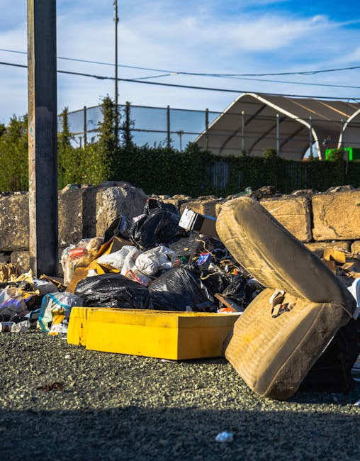 A large outdoor waste disposal area shows an overflowing collection of various rubbish items, including black bin bags, flattened cardboard boxes, and miscellaneous packaging materials, some spilling onto the paved ground. At the centre, a grey mixed paper and cardboard recycling bin is open, with its lid tilted back, revealing piled paper waste, such as newspapers, cardboard sheets, and paper bags. To the right, black waste bins are visible, one of which is filled with black plastic trash bags, while another red bin adjacent to them appears to be for general waste. A smaller white cardboard box and additional loose packaging are scattered at the front, with some items overlapping on the paved surface. Behind this scene, a blue and yellow commercial building with storefront signage and large windows is partially obscured by two parked cars—one grey on the left side of the image and another dark-colored. The background features a metal fence and an upper-floor balcony or scaffolding structure, suggesting ongoing building work or maintenance. The overall scene depicts a high volume of discarded waste awaiting collection, typical of private rubbish removal or alternative waste handling arrangements outside standard council services, with mild natural lighting indicating an overcast day.