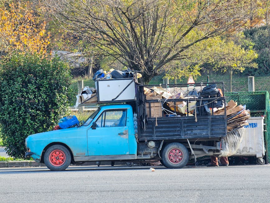 A light blue, compact flatbed truck parked on the side of a street, loaded with a large amount of mixed waste materials including flattened cardboard boxes, black plastic bags, wooden planks, and various discarded items. The truck's cab and body are painted in a matte light blue finish, with red-painted steel wheels. The cargo area is surrounded by a metal frame and is filled to capacity with waste, piled high and spilling over the sides. The environment includes a background of green hedges and a large deciduous tree with sparse, yellowing leaves, indicating autumn. The lighting is natural, suggesting daytime, with soft shadows cast on the street. The scene reflects an example of on-site waste collection, possibly by a private company like Waste Clearance Maida Vale, illustrating the kind of rubbish typically handled in non-public waste disposal services.