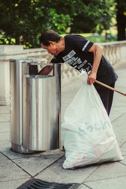 A man with short, dark hair and wearing a black T-shirt is seen depositing a white garbage bag into a cylindrical stainless steel public rubbish bin on a paved outdoor area. The bin has a brushed metal finish, with a slot at the top for waste disposal, positioned on the left side of the image. Behind him, there is a low concrete wall and lush green trees, suggesting a park or urban outdoor environment. The man appears to be engaged in waste disposal as part of an on-site rubbish collection or cleanup effort. The natural light indicates daytime, and the scene reflects a concern for cleanliness and waste management, aligning with private rubbish removal services such as waste clearance specialists like Waste Clearance Maida Vale. The setting emphasizes practical waste disposal, suitable for contextually supporting content about alternative rubbish collection methods outside local authority services.