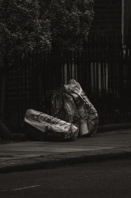 Three large black plastic rubbish bags, which appear to be full and somewhat crumpled, are placed on the pavement near a dark metal fence with vertical bars. One of the bags has visible white print on it, possibly indicating waste or recycling labels. The scene is set outdoors, adjacent to a residential area, with a concrete sidewalk and street surface in the foreground. Behind the fence, leafy trees or shrubs are visible, providing a natural backdrop. The lighting suggests an overcast or shaded environment, with subdued tones emphasizing the presence of waste awaiting collection or disposal. This scene relates to private or independent waste handling services, such as those provided by Waste Clearance Maida Vale, and highlights typical rubbish disposal practices outside urban properties, illustrating how waste is often temporarily stored on the street for clearance while adhering to local regulations.